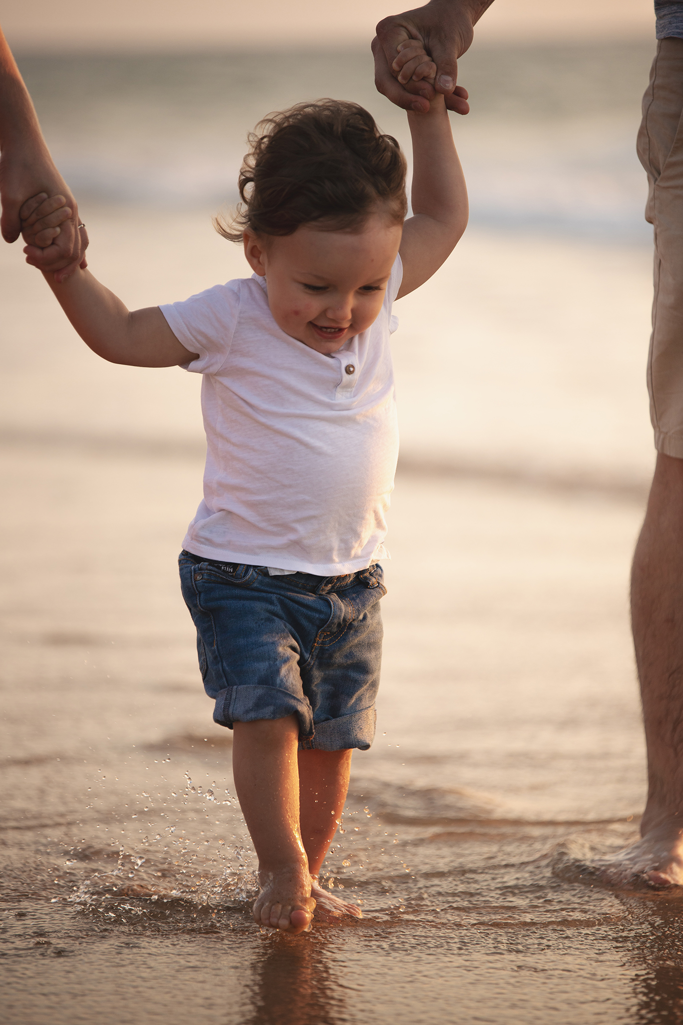Un enfant marche pieds nus sur la plage pendant la séance photo famille. Ses pieds trempent dans l'eau de l'océan. Il tient les mains de ses parents. Un souvenir créé par Meyline Esparcieux, photographe famille Dax, Landes.