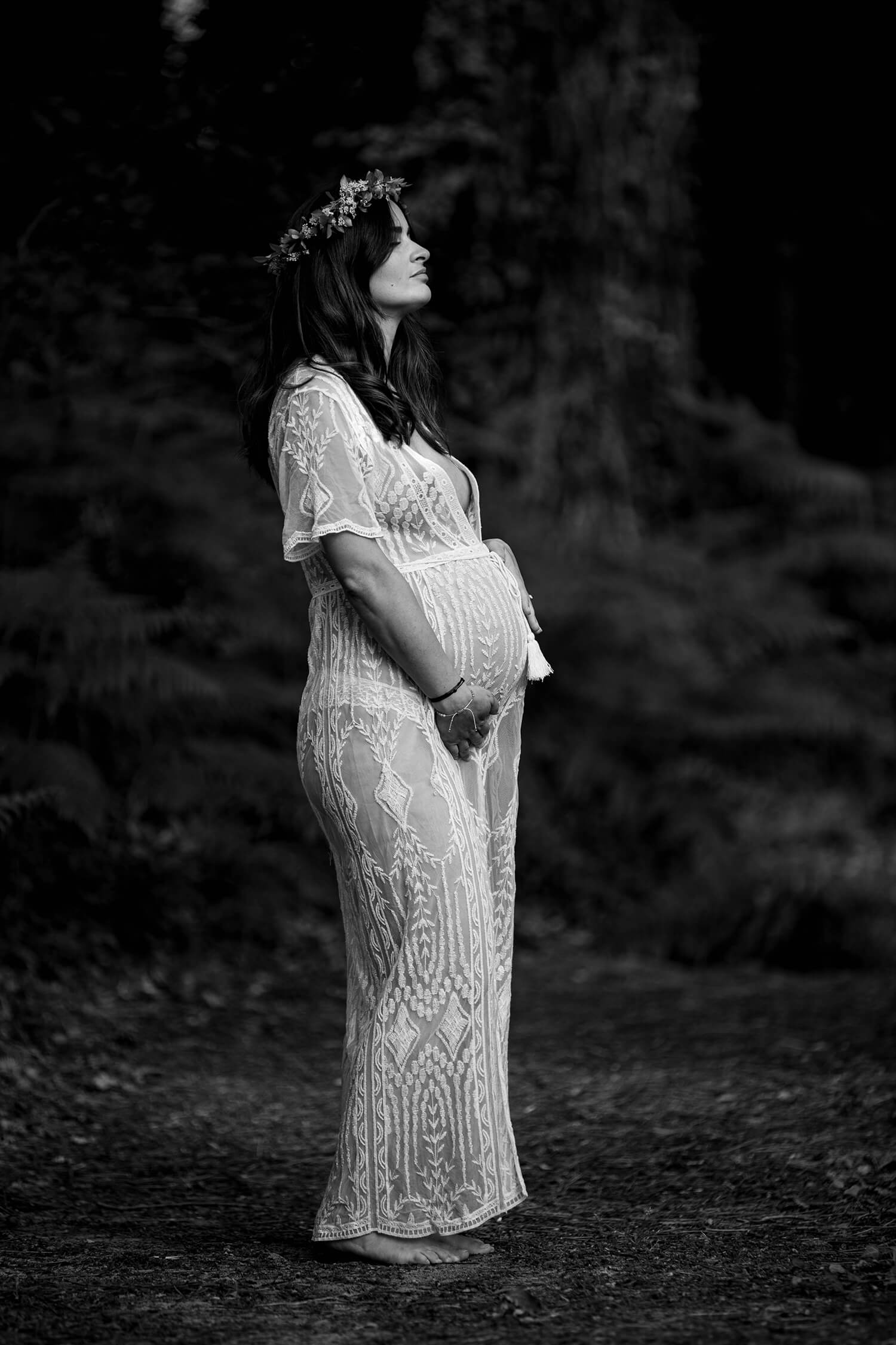 Une femme enceinte est habillée d'une robe longue bohème en dentelle et a une couronne de fleurs. Elle est debout dans la forêt pour sa séance photo grossesse. Une photo réalisée par Meyline Esparcieux, photographe grossesse Dax, Landes.