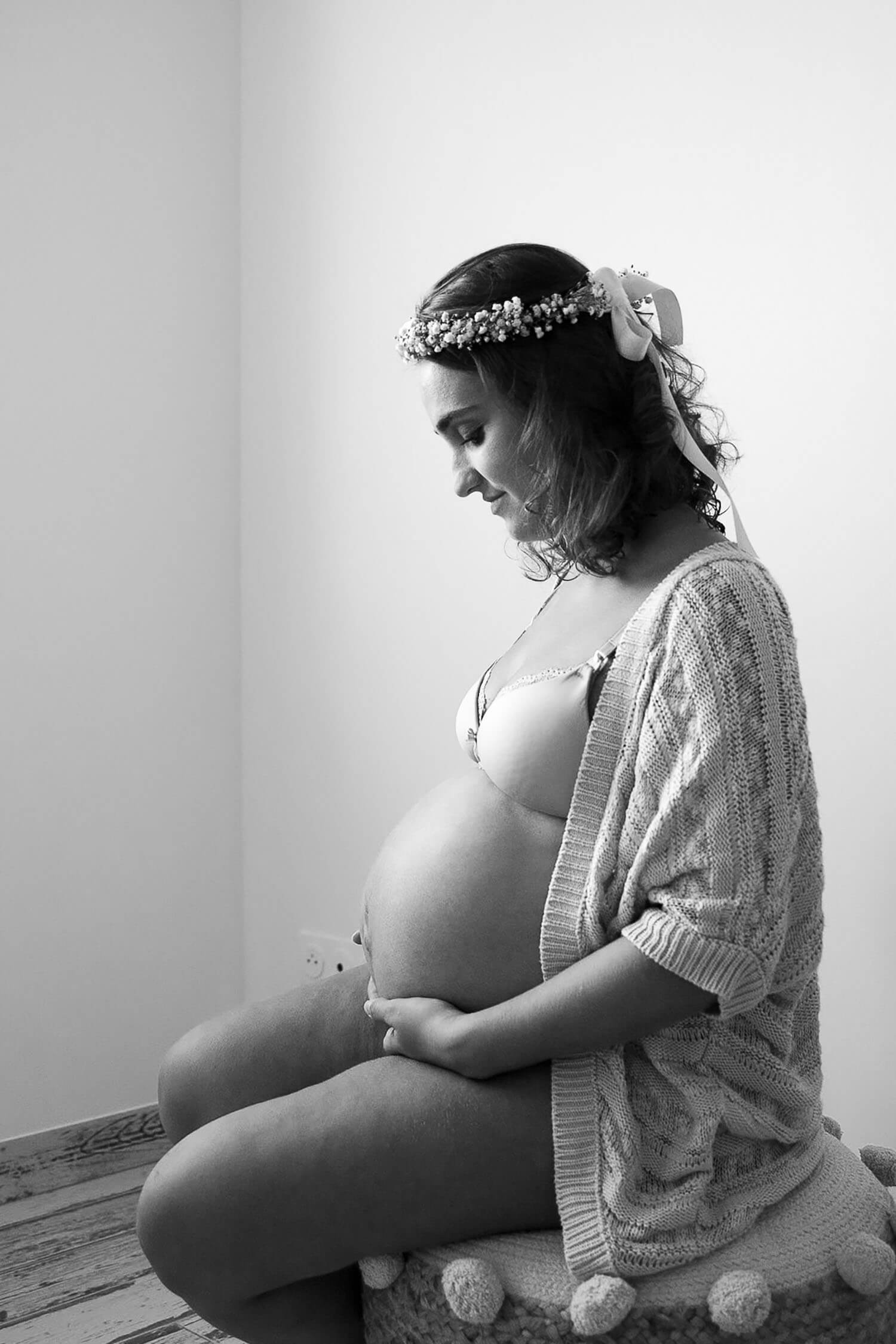 Une femme enceinte est assise de profil sur un tabouret. Elle touche son ventre rond et le regarde avec tendresse. Un moment tout doux capturé par Meyline Esparcieux, photographe grossesse Dax, Landes.