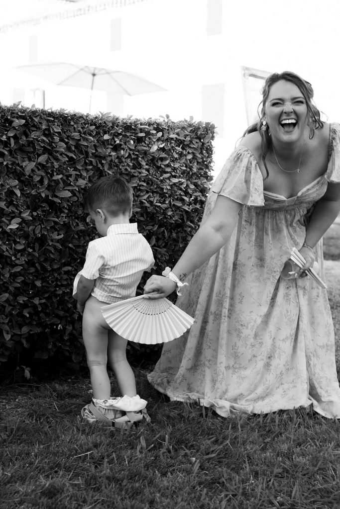 Un petit garçon d'honneur fait pipi contre une haie pendant la journée de mariage. Son pantalon est baissé. La mariée cache ses fesses avec son éventail et explose de rire. Un moment drôle capturé par Meyline Esparcieux, photographe reportage mariage Dax, Landes.