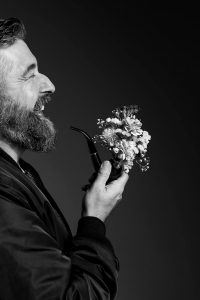 Portrait d'un homme barbu qui rit et tient une pipe décorée de fleurs. Une direction artistique par Meyline Esparcieux, photographe Dax.