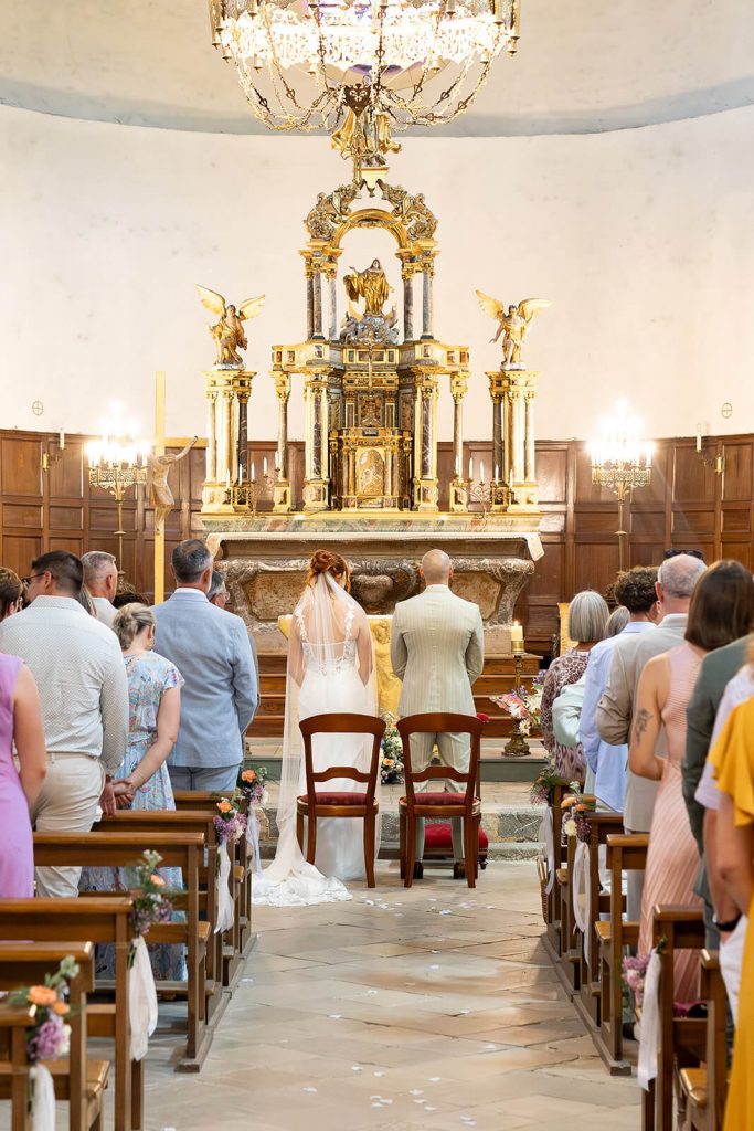 Cérémonie de mariage à l'église, on voit le couple de mariés debout devant l'autel, avec tous leurs proches derrière eux. Une photo de mariage créée par Meyline Esparcieux, photographe de mariage dans les Landes, basée à Dax.