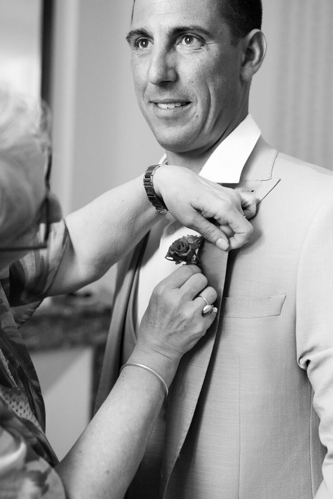 Le père du marié attache la boutonnière fleurie sur le costume du marié. Un moment des préparatifs par Meyline Esparcieux, photographe mariage Dax Landes.
