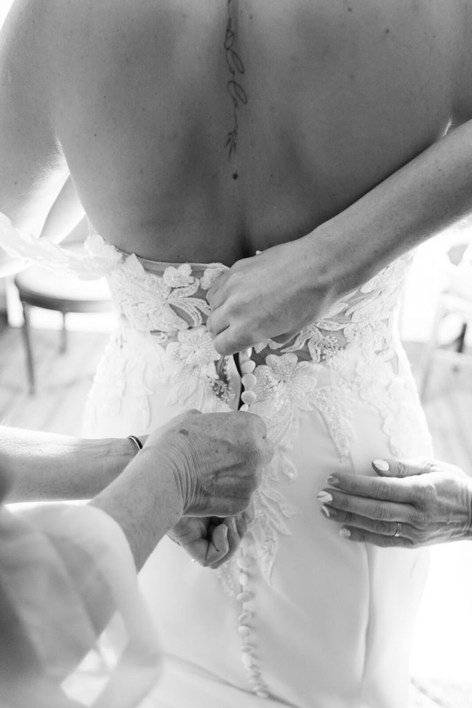 Les témoins aident la mariée à enfiler sa robe. Leurs mains ferment les boutons dans le dos de la mariée. Une photo de détail pendant les préparatifs de la mariée saisie par par Meyline Esparcieux, photographe mariage Dax Landes.